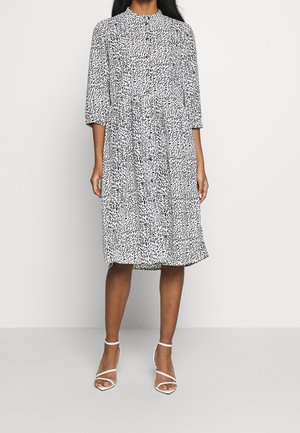 Robe à motif noir et blanc avec un col haut, des manches trois-quarts et une taille légèrement froncée, assortie à des talons blancs à lanières.