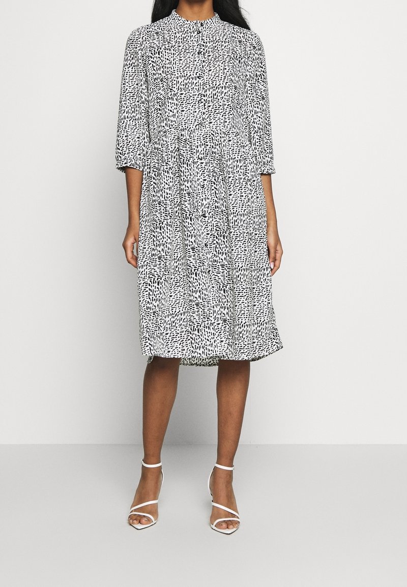 Abito a fantasia bianco e nero con scollo alto, maniche a tre quarti e una leggera vita arricciata, abbinato a tacchi bianchi con strappo.