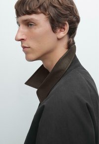 Black coat featuring a high, contrasting brown collar. The fabric appears smooth with a structured shape, shown from a side profile view.