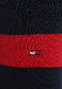 Calcetines a rayas en azul marino y rojo con textura acanalada. Presenta un pequeño logo en blanco, rojo y azul marino cerca del puño.