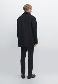 Cappotto di lana nera con taglio dritto, ampio colletto e spacchi laterali, abbinato a pantaloni neri slim-fit e stivaletti. Sfondo neutro.
