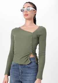 Långärmad, olivgrön, ribbad tröja med V-hals, sidoknappar och en liten öppning i fållen. Buren med högmidjade jeans.