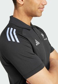 Polo nero con colletto, caratterizzato da accenti bianchi a tre strisce sulle spalle e un logo sul petto. Realizzato in tessuto morbido.