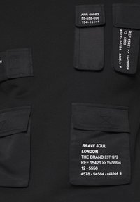 Černá textilie s několika kapsami na záplaty. Každá kapsa má bílý text včetně kódů a značkových detailů. Hladká textura látky.