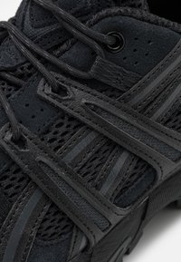 Schwarzer Sportschuh mit einem Obermaterial aus Mesh und synthetischem Material, strukturiertem Oberflächendesign, kontrastierenden glatten Akzenten und einem ausgeklügelten Schnürsystem.