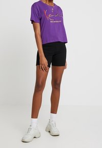 Lila kortärmad t-shirt med broderad text, ihopparat med svarta shorts. Vita sneakers med strukturerade sulor och synliga snören.