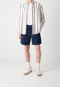 Bílá a navy proužkovaná košile s knoflíky, pod ní bílý tričko, tmavě modré šortky, bílé tenisky s hnědými akcenty a bílé kotníkové ponožky.