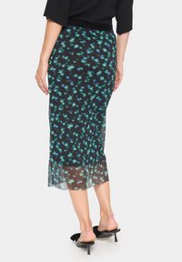 Jupe midi plissée en tissu noir transparent avec imprimé floral vert et bleu. Elle présente une taille ajustée et un détail fente sur le côté. Portée avec des chaussures à talons noires.
