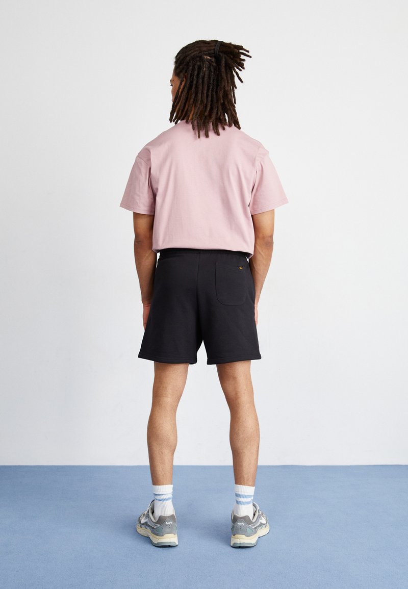 Hellrosa Baumwoll-T-Shirt, getragen mit schwarzen Shorts, die eine Gesäßtasche haben. Graue Sneaker mit blau-weißen gestreiften Socken runden das Outfit ab.