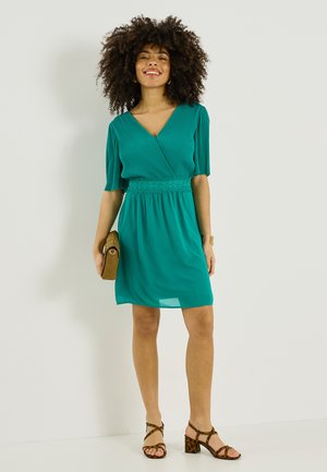 Mujer de cabello rizado con un vestido azul verdoso hasta la rodilla, tacones marrones con estampado de leopardo, sosteniendo un bolso de mano marrón, sonriendo frente a un fondo liso.