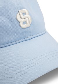 Lyseblå baseballcap laget av teksturert stoff. Har et hvitt brodert logo og synlige sømmer langs skyggen.