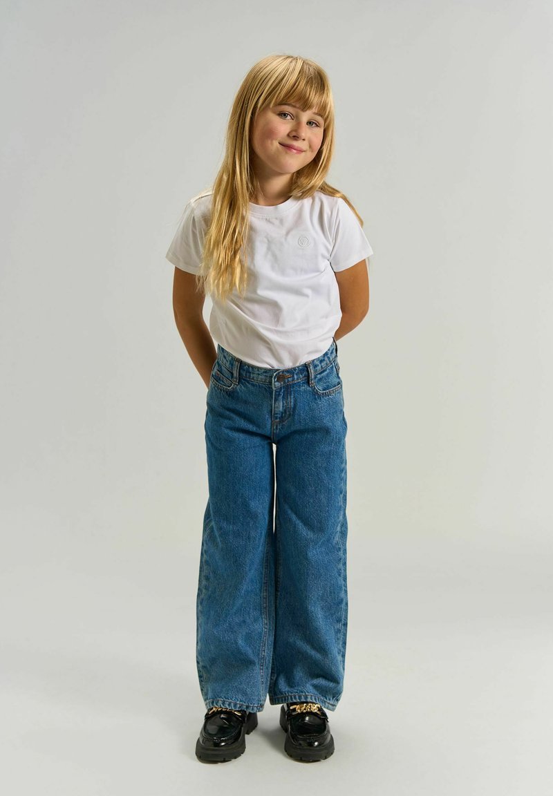 Jeune fille aux longs cheveux blonds se tient en souriant, portant un t-shirt blanc, un jean bleu à jambes larges et des chaussures noires avec un détail chaîne dorée.