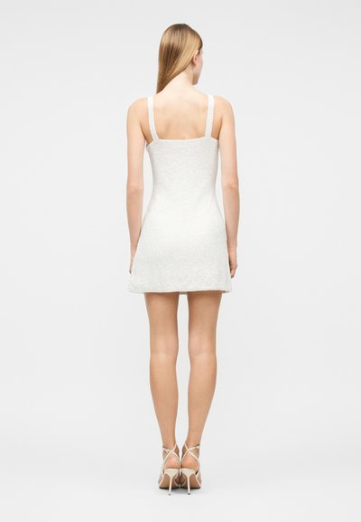 Γυναίκα με μακριά ξανθά μαλλιά που φοράει λευκό υφασμάτινο μίνι φόρεμα χωρίς μανίκια και μπεζ πέδιλα με ψηλό τακούνι, στέκεται γυρισμένη με την πλάτη.