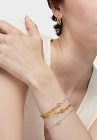 Braccialetti d'oro e d'argento con disegni a catena e dettagli rotondi. Il modello indossa un orecchino a cerchio d'oro; il tono della pelle è visibilmente chiaro.