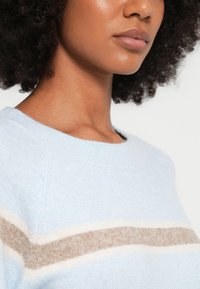 Moteris su garbanotais plaukais dėvi šviesiai mėlyną megztinį su horizontalia smėlio spalvos ir baltos spalvos juosta per krūtinę.