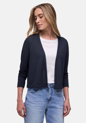 Mujer que lleva un cárdigan abierto de color azul marino oscuro sobre una camiseta blanca y jeans azul claro, mirando ligeramente hacia abajo a su izquierda.