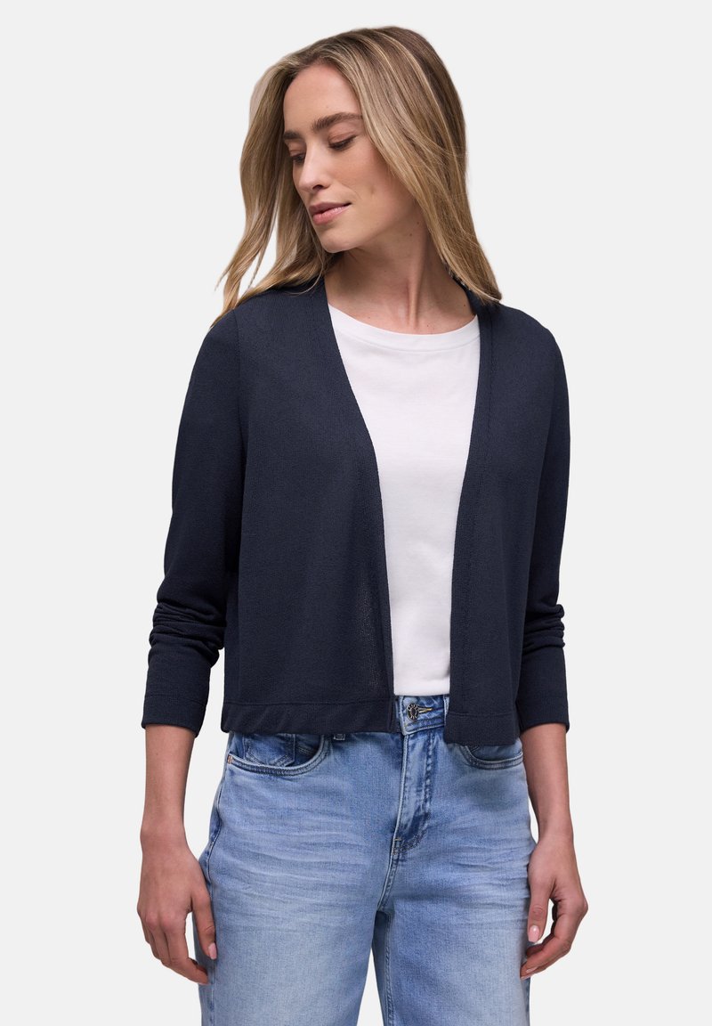 Mujer que lleva un cárdigan abierto de color azul marino oscuro sobre una camiseta blanca y jeans azul claro, mirando ligeramente hacia abajo a su izquierda.