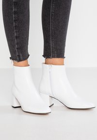 Bottines blanches à finition en cuir lisse, talons carrés et fermeture éclair latérale, assorties à un jean skinny noir à ourlet effiloché.