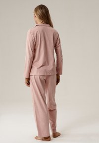 Kobieta stojąca boso z długimi włosami, ubrana w jasnoróżowy zestaw piżamowy z delikatnym wzorem księżyca i gwiazd, zwrócona plecami w stronę jednolitego tła.