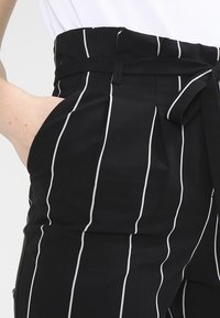 Černé kalhoty s vysokým pasem a bílými vertikálními pruhy, které mají zavázaný pásek, boční kapsy a hladkou texturu látky.