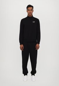 Sort zip-up sweatshirt med høj krave, matchende joggers og hvide sneakers. Har EA7-logo på brystet. Glat tekstur.