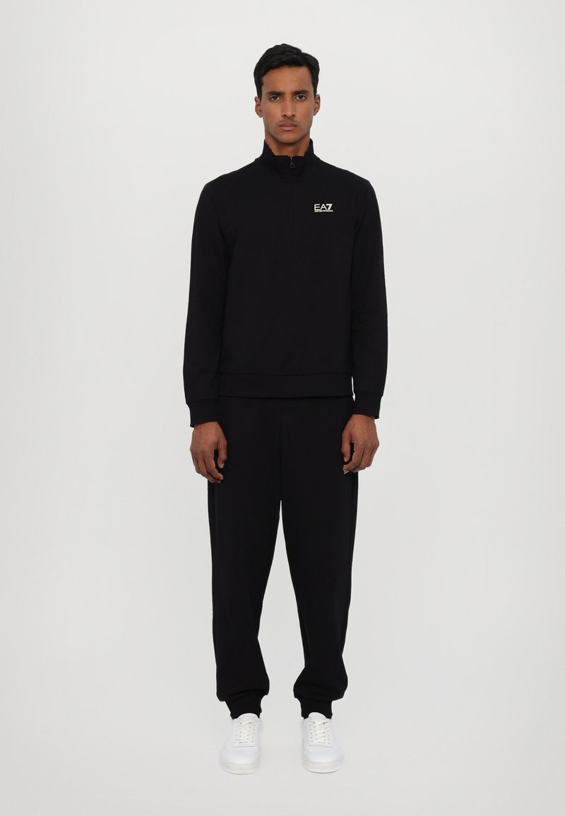 Sort zip-up sweatshirt med høj krave, matchende joggers og hvide sneakers. Har EA7-logo på brystet. Glat tekstur.
