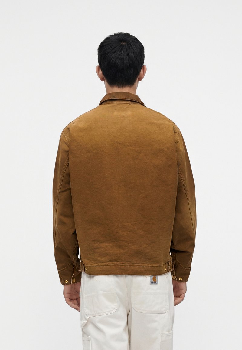 Mann trägt braune Jacke und weiße Hose, von hinten vor einem einfarbigen hellen Hintergrund gezeigt.