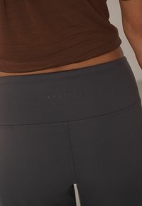 Aktīvā apģērba legingi tumši pelēkā audumā, ar gludu tekstūru, augstu jostasvietu un diskrētu zīmolu aizmugurē.