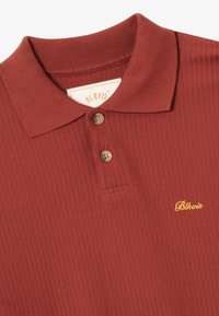 Prążkowana, rdzawego koloru koszulka polo z dwoma guzikami i żółtym haftem "Blkvis" na lewej piersi.