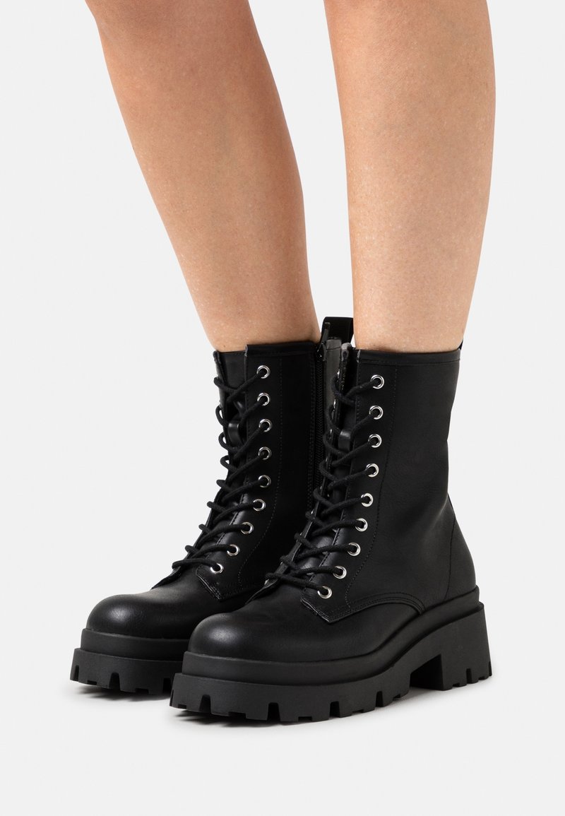 Bottes de combat noires à lacets avec une semelle épaisse, dotées d'œillets argentés et d'une fermeture éclair latérale. Construites en cuir lisse à texture mate.
