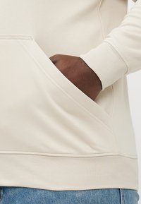 Beige hoodie met een kangoeroezak, ribgebreide manchetten en zoom. Het materiaal is zacht met een gladde textuur; het heeft een eenvoudige, minimalistische uitstraling.