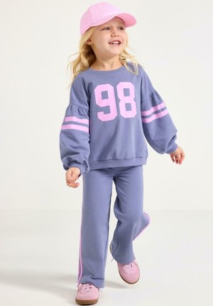 Lyse lilla sweatshirt med lyserødt nummer "98" og stribede ærmer, kombineret med matchende bukser med lyserøde sidestriber og lyserøde sneakers.