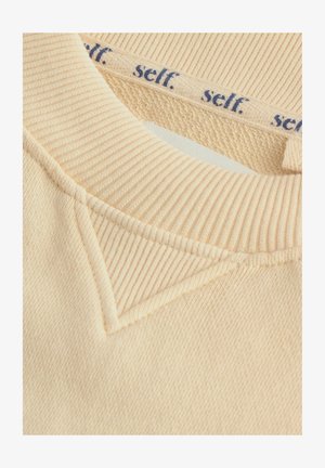 Beige Pulloverkragen mit gerippter Textur und einem Baumwollgefühl. Enthält ein Etikett mit "self." in Marineblau gedruckt. Detaillierte Nahtakzente sind sichtbar.