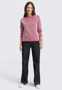 Różowy sweter w paski z dzianiny, w połączeniu z granatowymi dżinsami. Prążkowana tekstura, okrągły dekolt i kontrastowe mankiety. Model ma na sobie białe trampki.