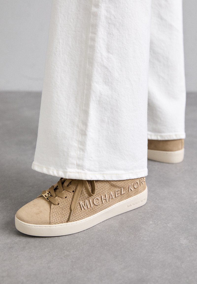 Beige Sneakers mit einem perforierten Wildleder-Obermaterial, flachen Schnürsenkeln und geprägtem Branding an der Seite. Kombiniert mit weißen, weiten Hosen.
