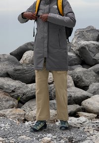 Szara wodoodporna kurtka z wysokim kołnierzem, beżowe spodnie cargo i solidne buty trekkingowe. Dostosowuje kurtkę w pobliżu skalistego terenu.