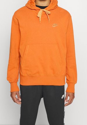 Mężczyzna ubrany w pomarańczową bluzę z kapturem Nike z troczkami i kieszenią kangurka, połączoną z czarnymi spodniami dresowymi Nike z białymi logotypami i bocznymi paskami.