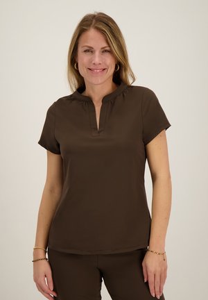 Vrouw met lichtbruin haar die gouden oorringen draagt, een donkerbruine korte mouw top met V-hals en bijpassende broek, glimlacht zacht.