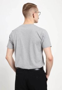 Muž s krátkými vlasy a brýlemi, oblečený v jednoduchém šedém tričku a černých kalhotách, zobrazený zezadu na bílém pozadí.