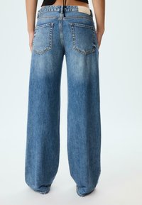 Široke modre denim hlače z zbledelim izgledom, ki imajo pet žepov, oznako na pasu in sproščen kroj, ki sega do gležnjev.