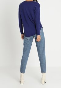 Granatowa bluzka z długim rękawem o luźnym kroju, zestawiona z jasnoniebieskimi dżinsami. Modelka ma na sobie kremowe botki na wysokim obcasie.