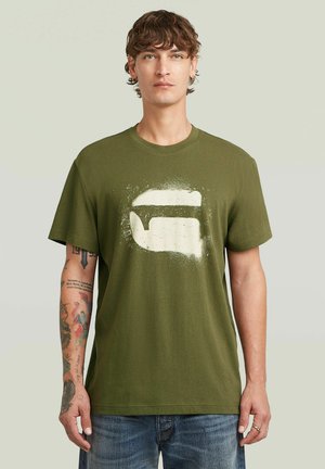 Joven con cabello rizado, vistiendo una camiseta verde oliva con un gráfico en estilo de pintura en aerosol blanco y jeans azules, de pie contra un fondo liso.