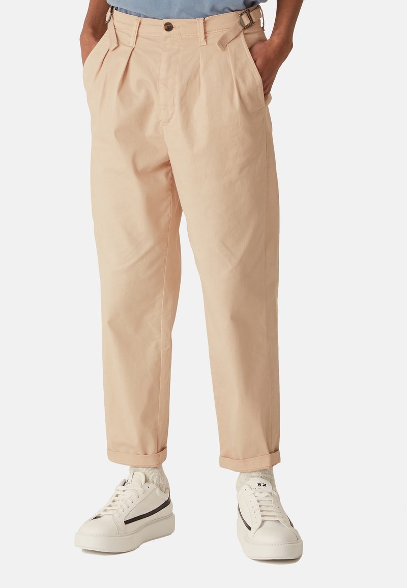 Spodní část těla osoby oblečené v béžových skládacích kalhotách s pásem, bílých teniskách a světle šedých ponožkách na bílém pozadí.