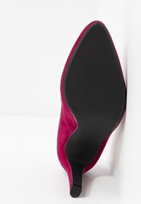 Chaussure à talon haut à bout pointu en daim bordeaux avec semelle noire texturée et talon fin, positionnée debout sur fond blanc.