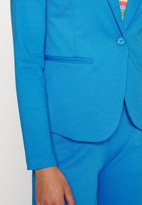 Jasně modrý ušitý blázr s jedním knoflíkem, slim fit a hladkým povrchem. Obsahuje jemný detail kapsy a odpovídající kalhoty.