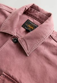 Close-up of a pink jacket collar with white stitching, a black button, and a black and orange brand label inside the collar.