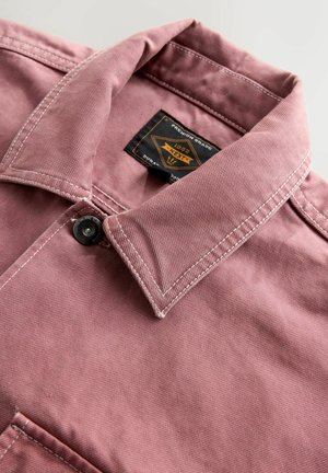 Nahaufnahme eines pinken Jackenkragens mit weißem Nähen, einem schwarzen Knopf und einem schwarzen und orangefarbenen Markenetikett im Kragen.