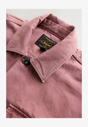 Artimu atvaizdu rodomas rausvos striukės apykaklė su baltu siūlu, juodu mygtuku ir juoda bei oranžine prekės ženklo etikete apykaklėje.