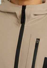 Felpa beige con cerniera frontale, dettagli in cerniera neri e una tasca sul lato sinistro. Tessuto liscio senza motivi visibili.