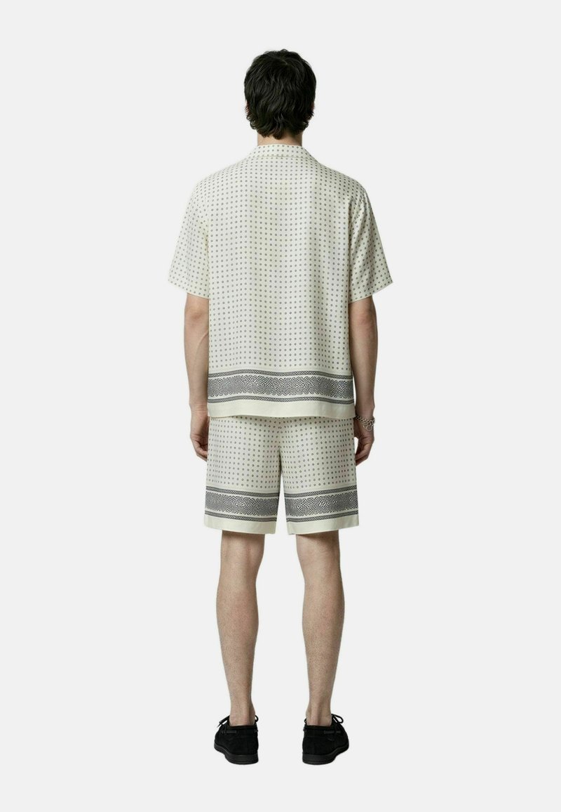 Hombre de espaldas, vistiendo una camisa de manga corta y pantalones cortos a juego con un pequeño estampado geométrico y zapatos negros.